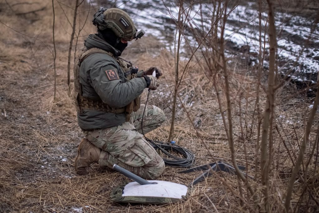 A helmeted soldier kneels among bare trees, handling a cable beside a coil of wire and a white satellite terminal.