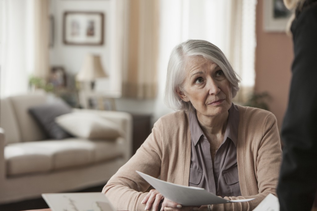 A gray-haired woman seated at a table holds papers and looks up at a person standing beside her.