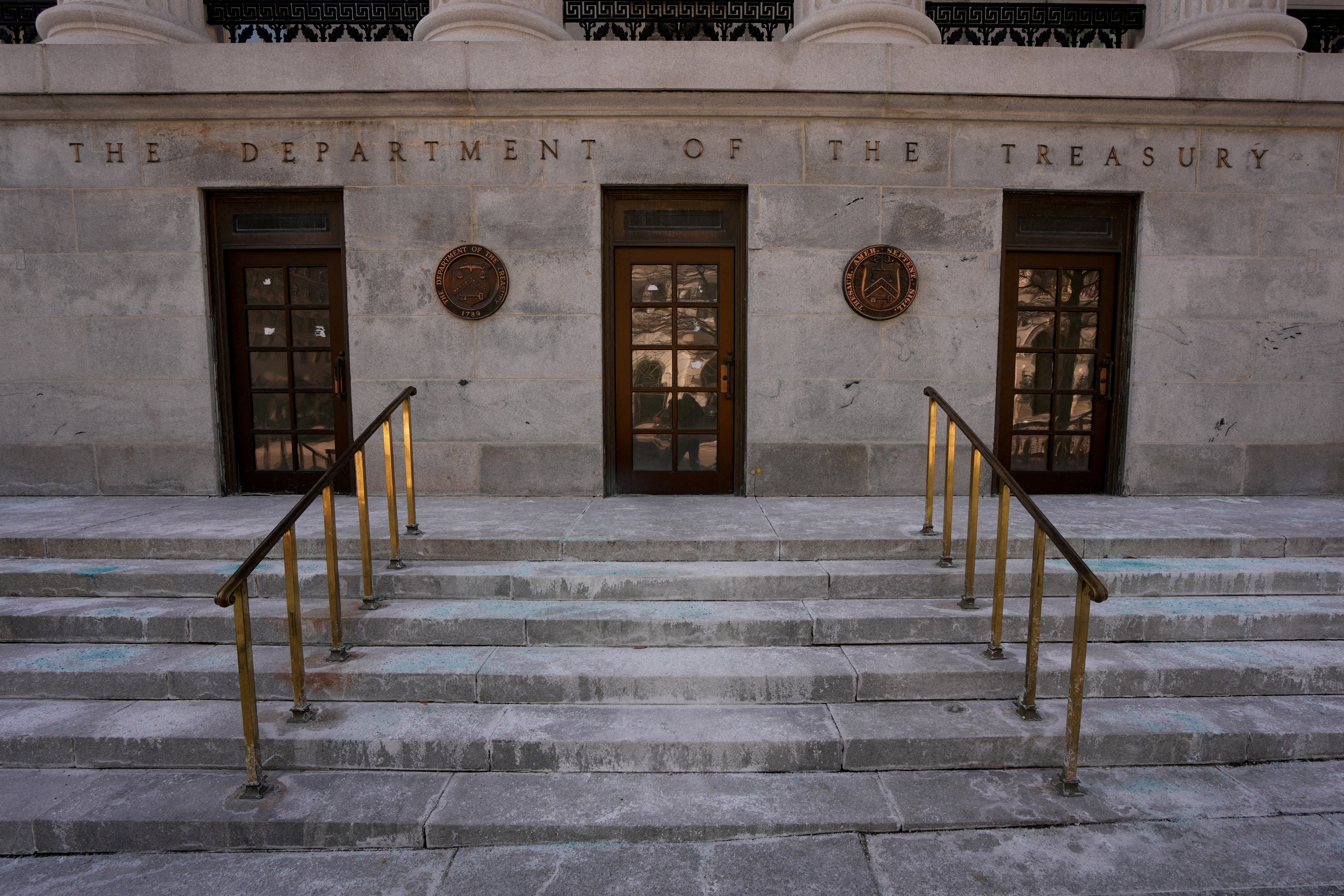The words The Department of the Treasury carved in stone above three wooden doors and two bronze seals, with brass handrails running down the granite steps.