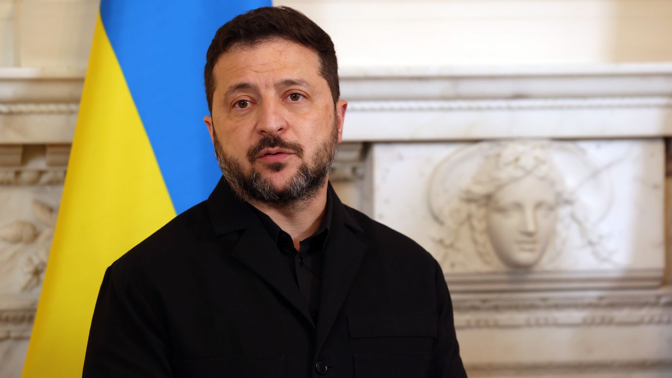 Volodymyr Zelensky, in a black jacket, stands beside a Ukrainian flag in front of a carved stone relief.