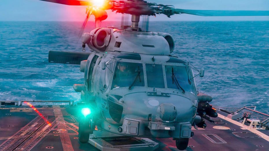 A gray military helicopter on a ship’s deck at sea, its rotor turning and red and green lights glowing.