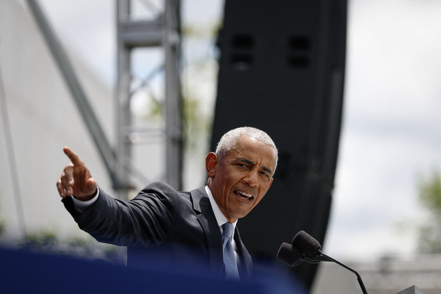 Barack Obama, at an outdoor lectern beside a microphone, points out to his right with his index finger, a speaker tower behind him.