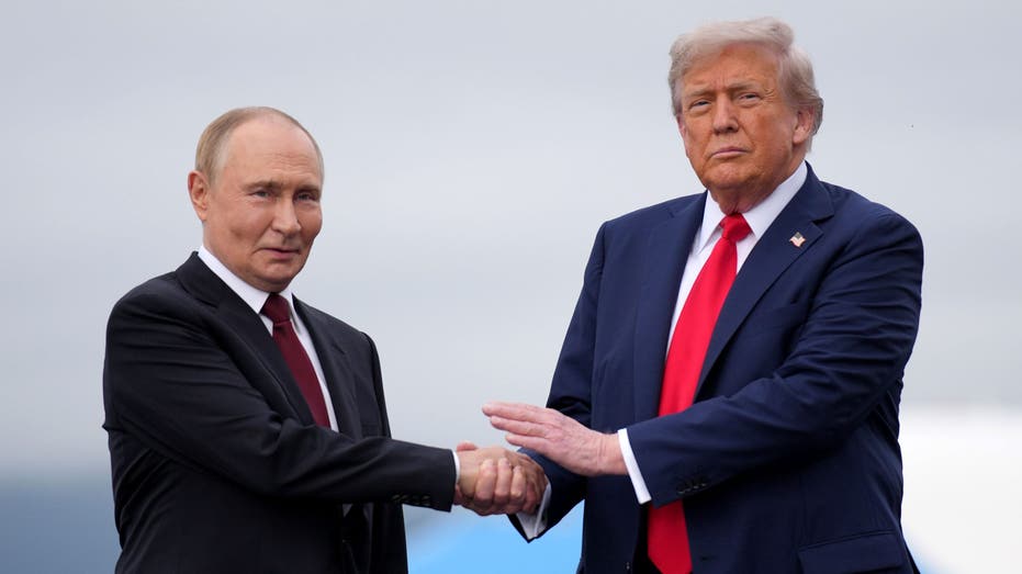 Trump shakes hands with another man in a dark suit while holding his other hand above their clasped hands.