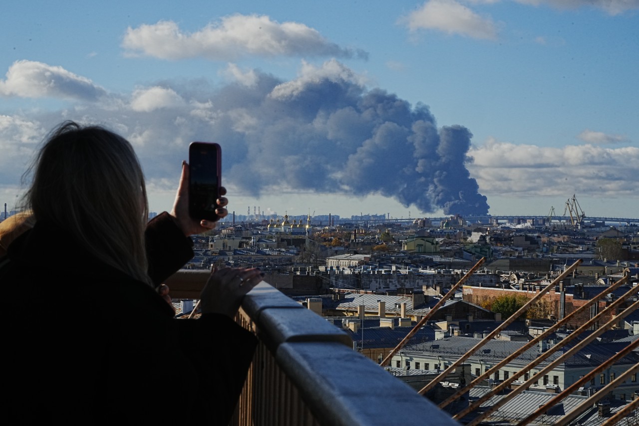 A woman on a rooftop holds up a phone toward a dark plume of smoke rising over the St. Petersburg skyline.
