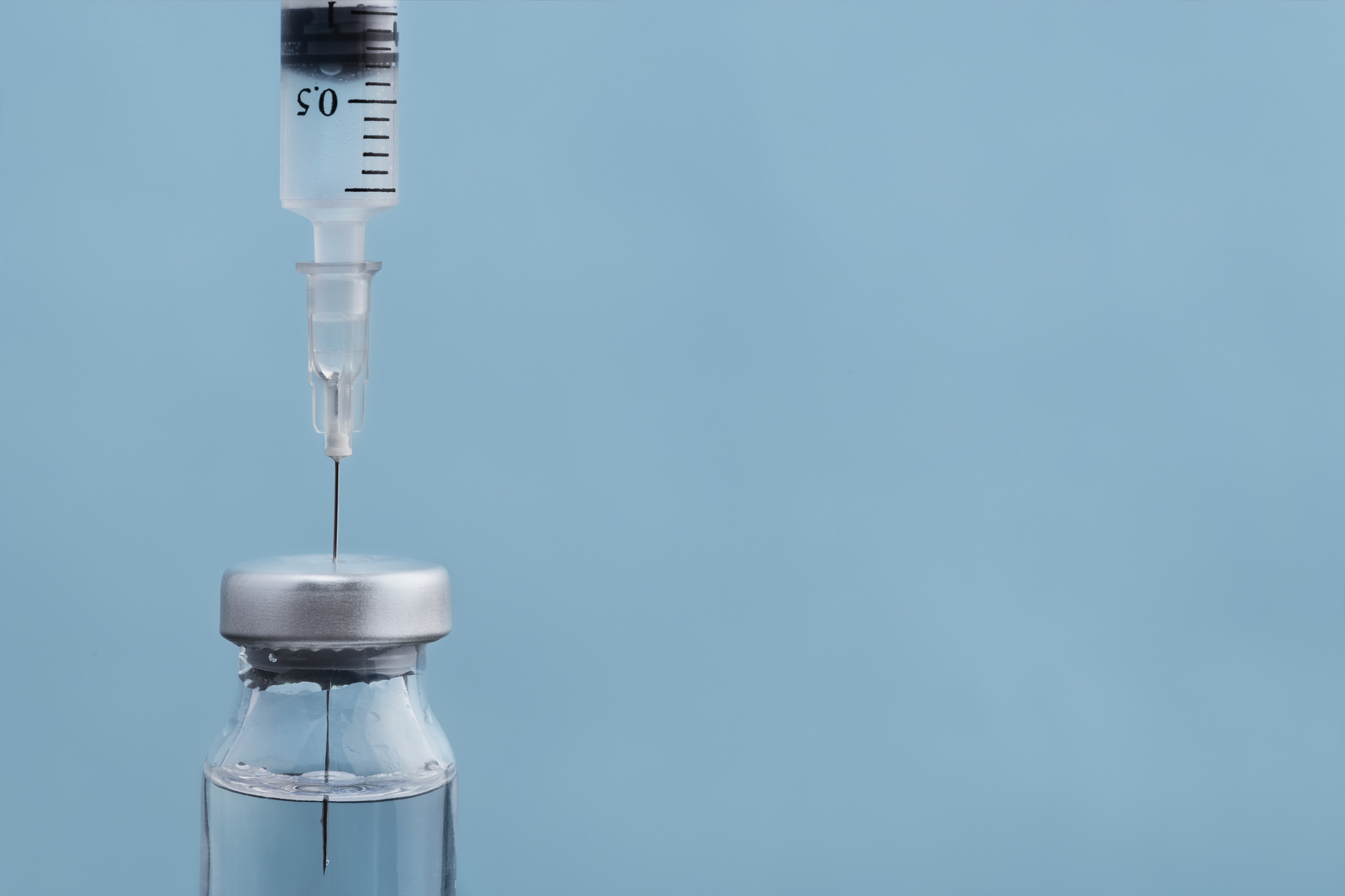A syringe needle pierces the silver cap of a small glass vial of clear liquid against a pale blue background.