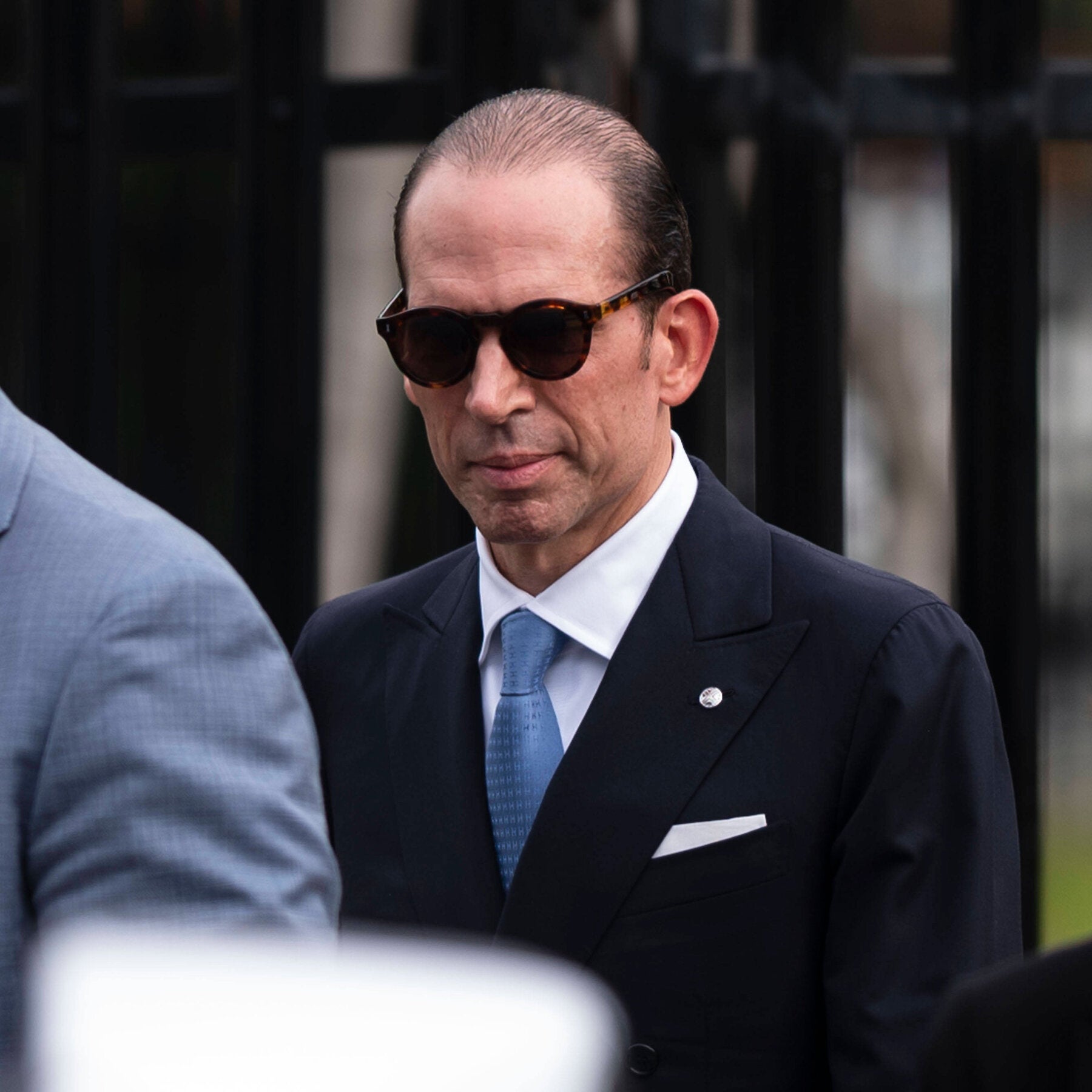 Julio Herrera Velutini, in dark sunglasses, a navy suit and a blue tie, stands in front of a black metal fence.