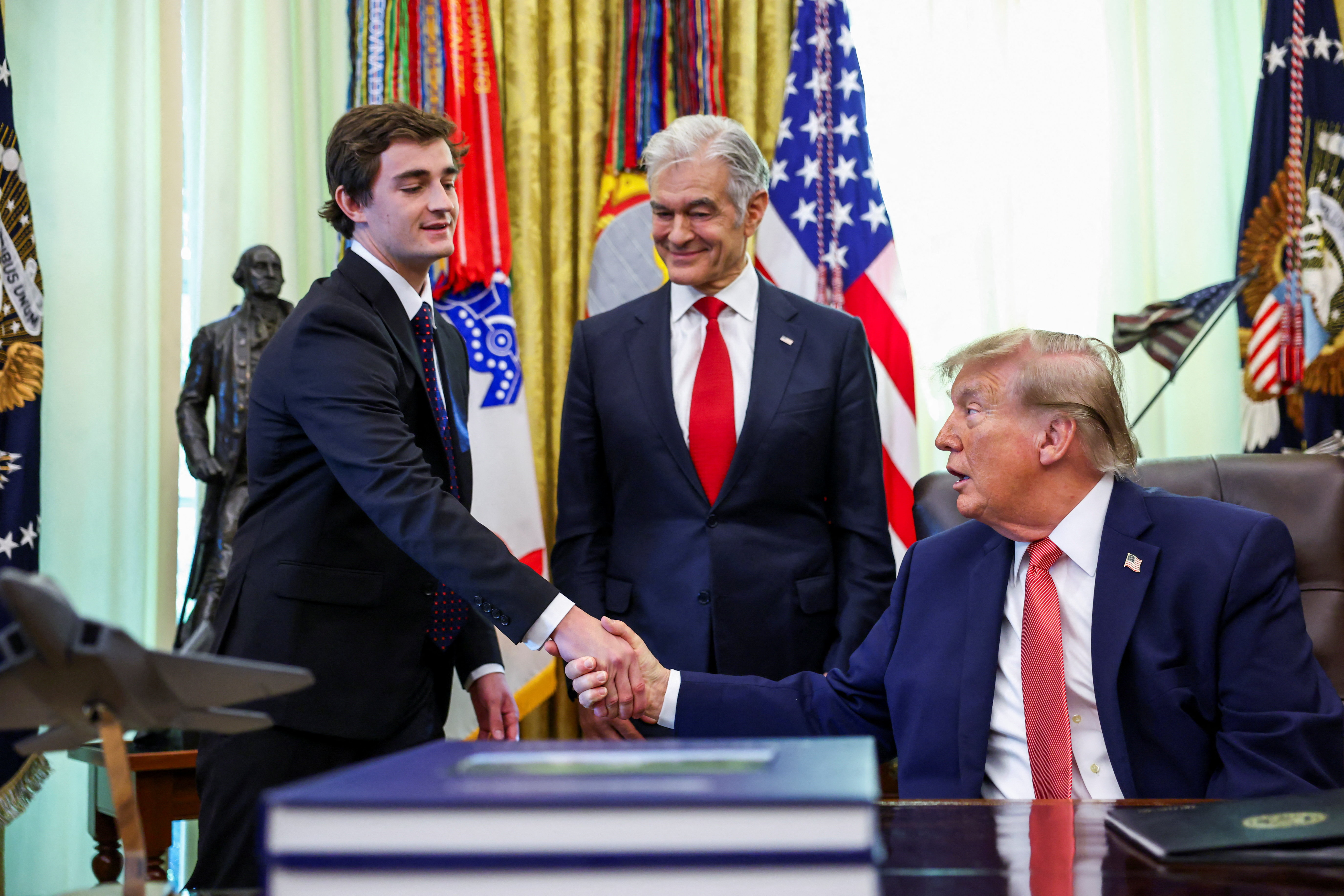 Trump, seated at his desk, shakes hands with a young man in a dark suit as another man stands between them in front of a row of flags.