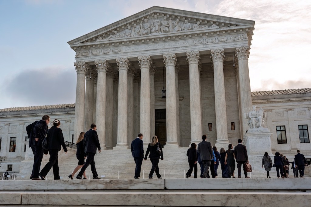 People in dark clothes climb the steps of the Supreme Court building under a pale evening sky.