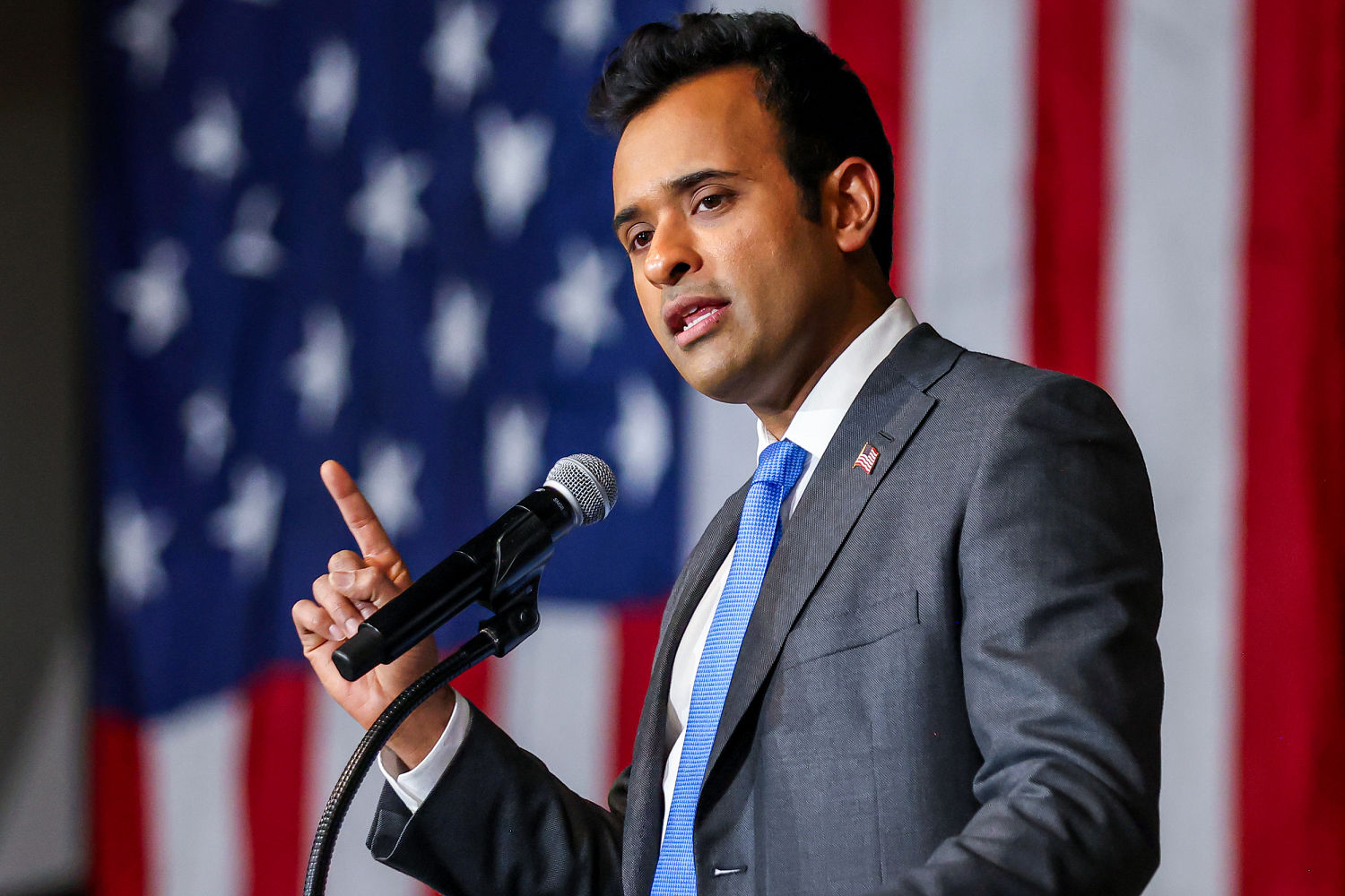 Ramaswamy raises an index finger while speaking into a microphone in front of an American flag.