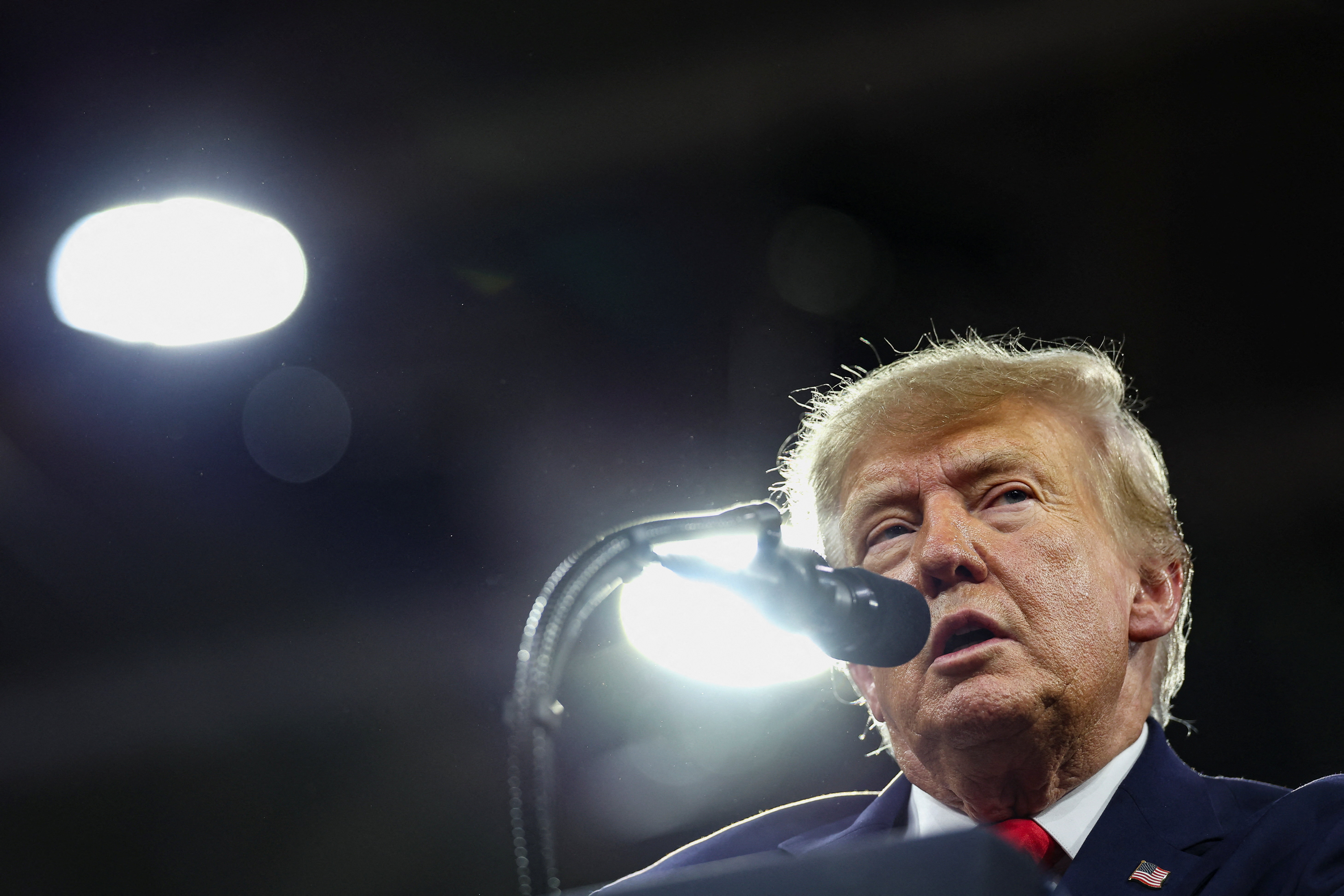 Trump speaks into a microphone under bright lights, his face lit from one side against a dark hall.