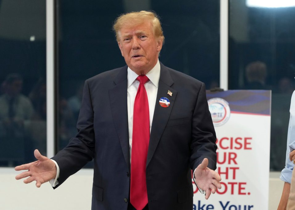 Trump stands with his hands spread apart, wearing a dark suit, red tie and round voting sticker.