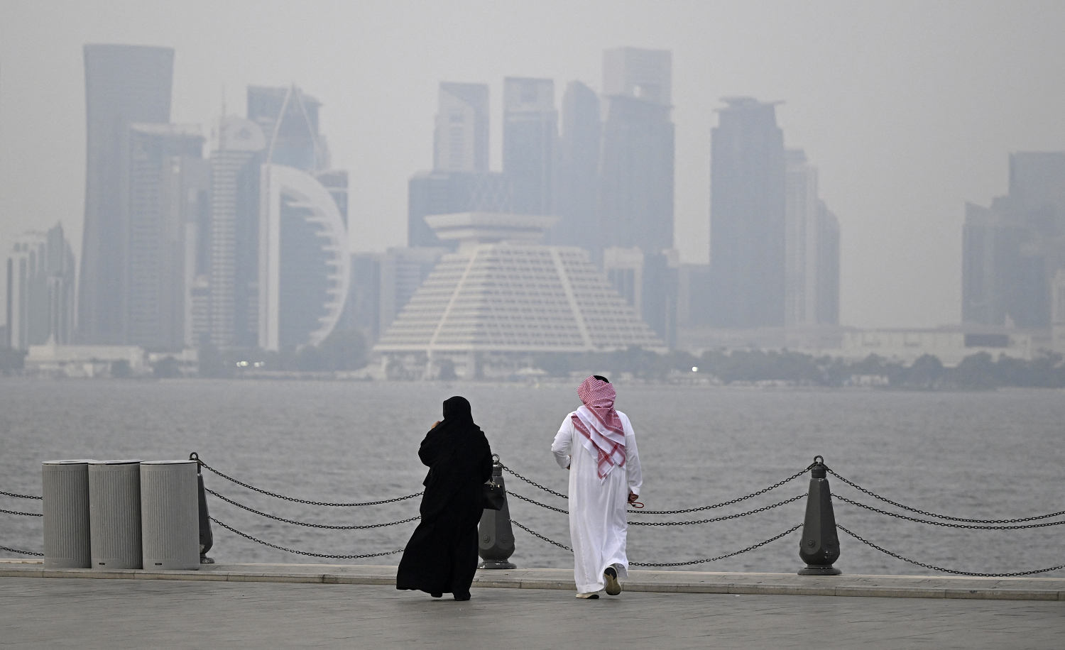 Two people in traditional clothing walk along a waterfront promenade, with a hazy skyline of high-rise buildings across the water.