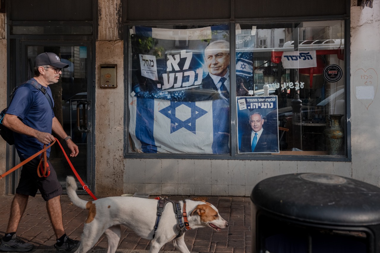 A man walks a dog past a storefront window displaying Israeli flags and posters featuring Netanyahu.