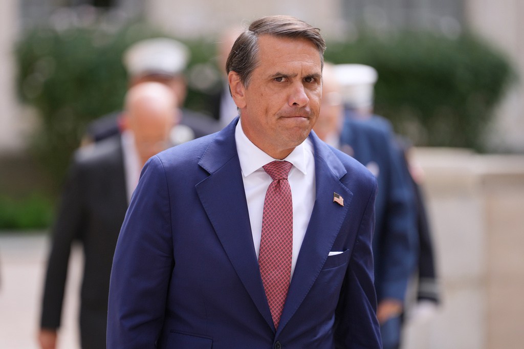 Blanche walks outdoors wearing a navy suit, patterned red tie, and American flag lapel pin, with several people behind him.