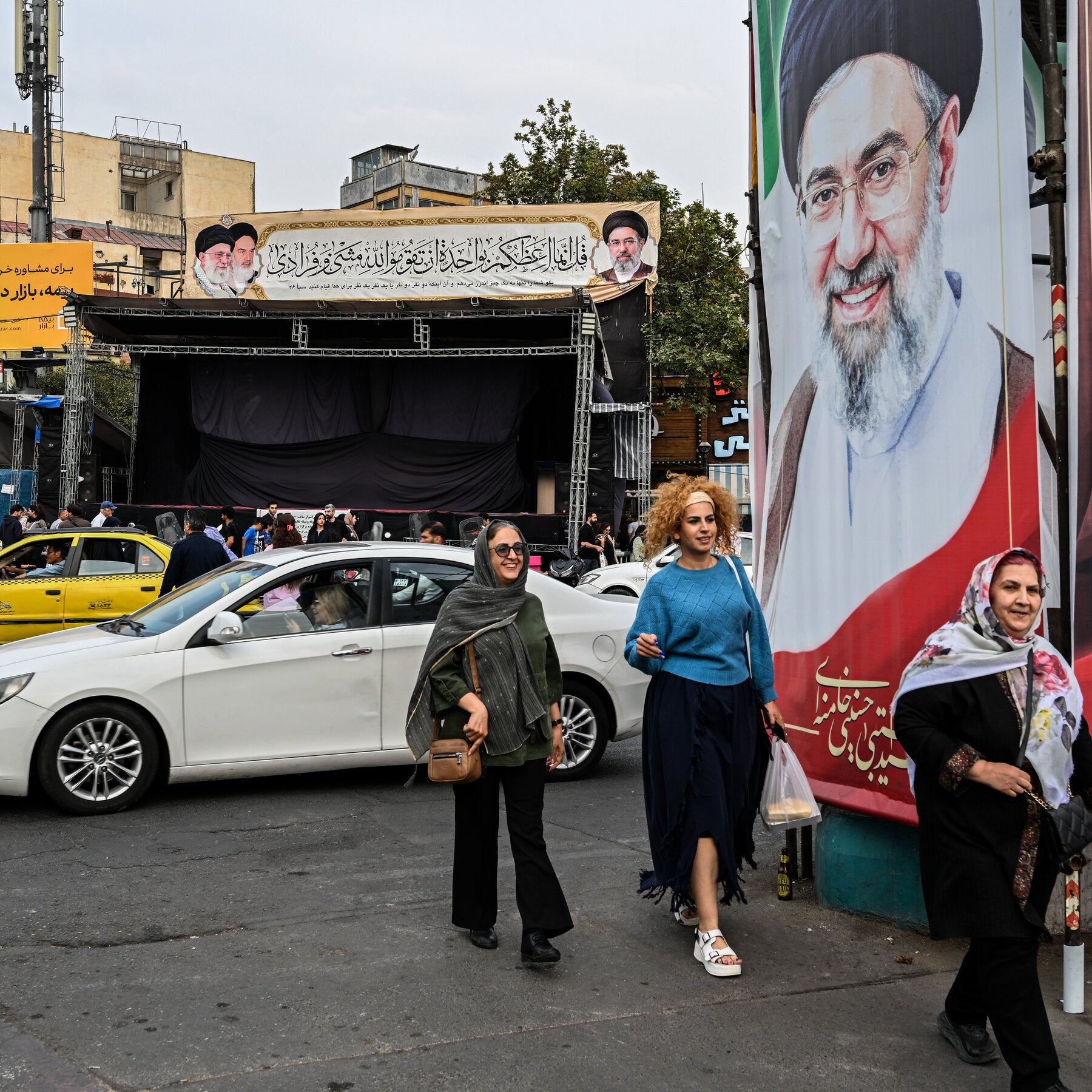 Women walk past cars and a large portrait of a cleric, with a black-draped stage and portrait banner behind them.