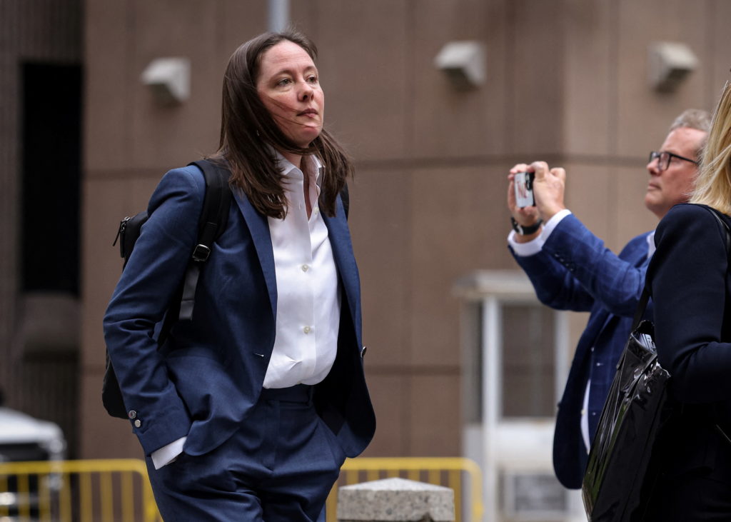 Maurene Comey walks outdoors in a navy suit with a backpack as a man nearby holds up a phone to photograph her.