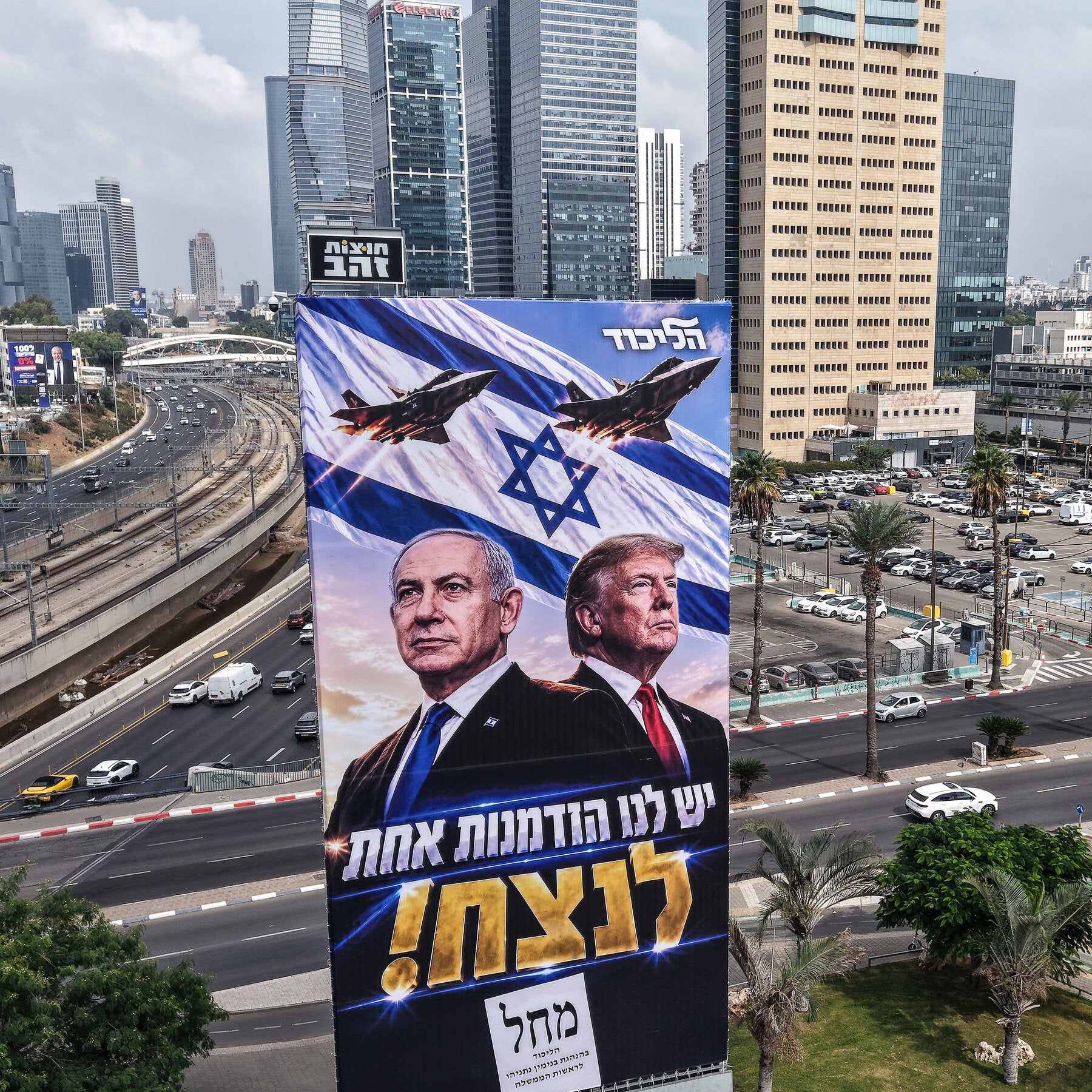 A towering billboard shows Netanyahu and another man in a suit beneath two fighter jets and an Israeli flag, above a highway and office towers.