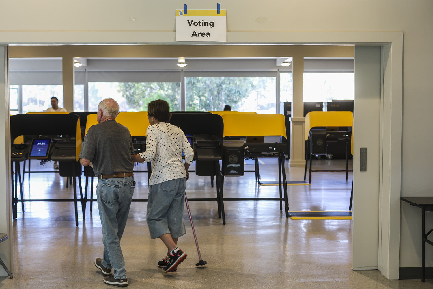 Two older people, one with a cane, walk arm in arm under a “Voting Area” sign toward rows of yellow voting booths.