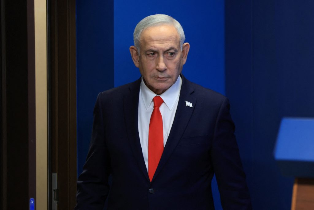 Netanyahu wears a dark suit and red tie while standing beside a doorway against a blue backdrop.