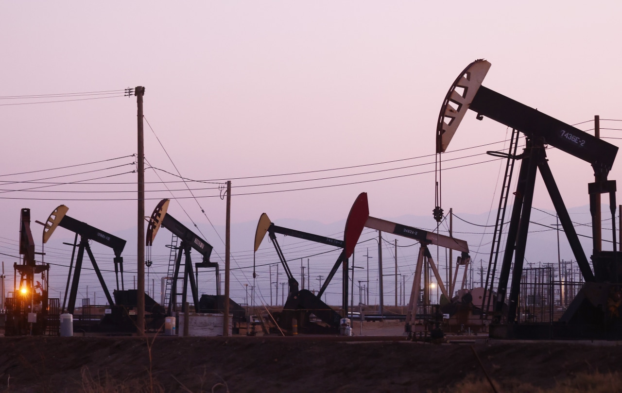 Several oil pumpjacks stand among utility poles and power lines beneath a pink sky.