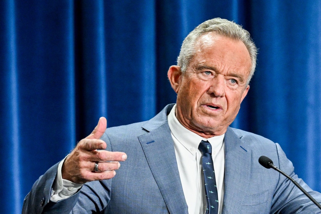 A gray-haired man in a light blue suit and patterned tie gestures beside a microphone against a blue curtain.