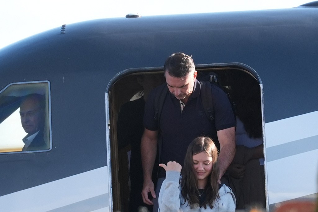 A man wearing a dark polo shirt and backpack stands in an aircraft doorway behind a girl in a gray sweatshirt.