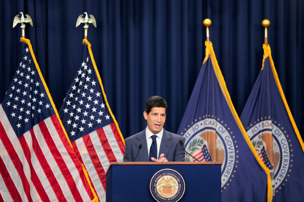 A man in a dark suit speaks at a Federal Reserve Board of Governors lectern between U.S. flags and two Board flags.
