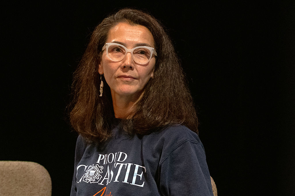 A woman in white-framed glasses and a navy “Proud Coastie” sweatshirt sits on a stage against a black backdrop.