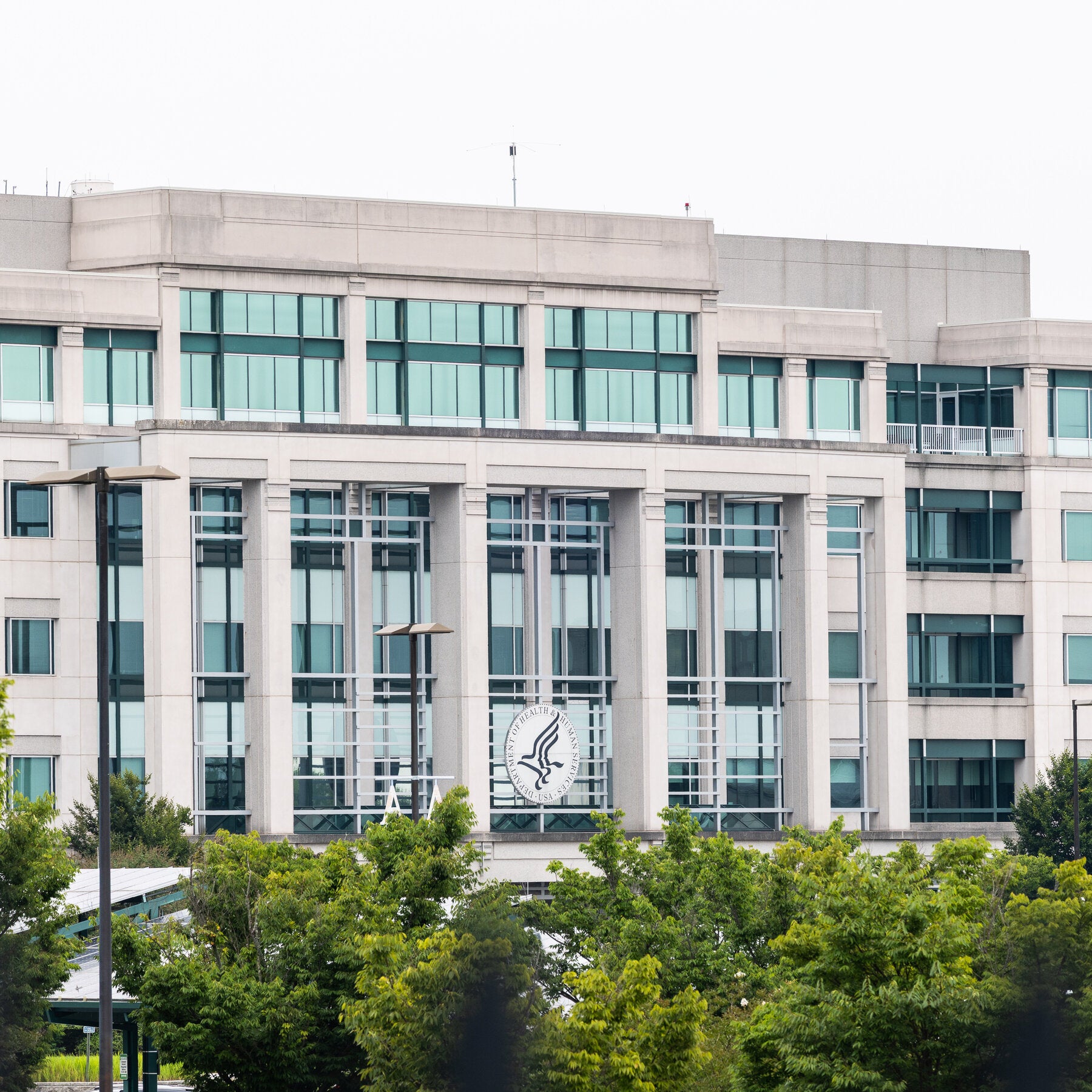 An office building with a Department of Health and Human Services seal on its facade, behind a row of trees.