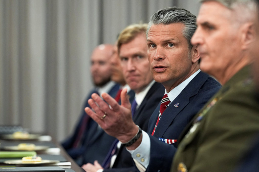 Hegseth gestures while seated at a meeting table with suited men and a uniformed military officer.