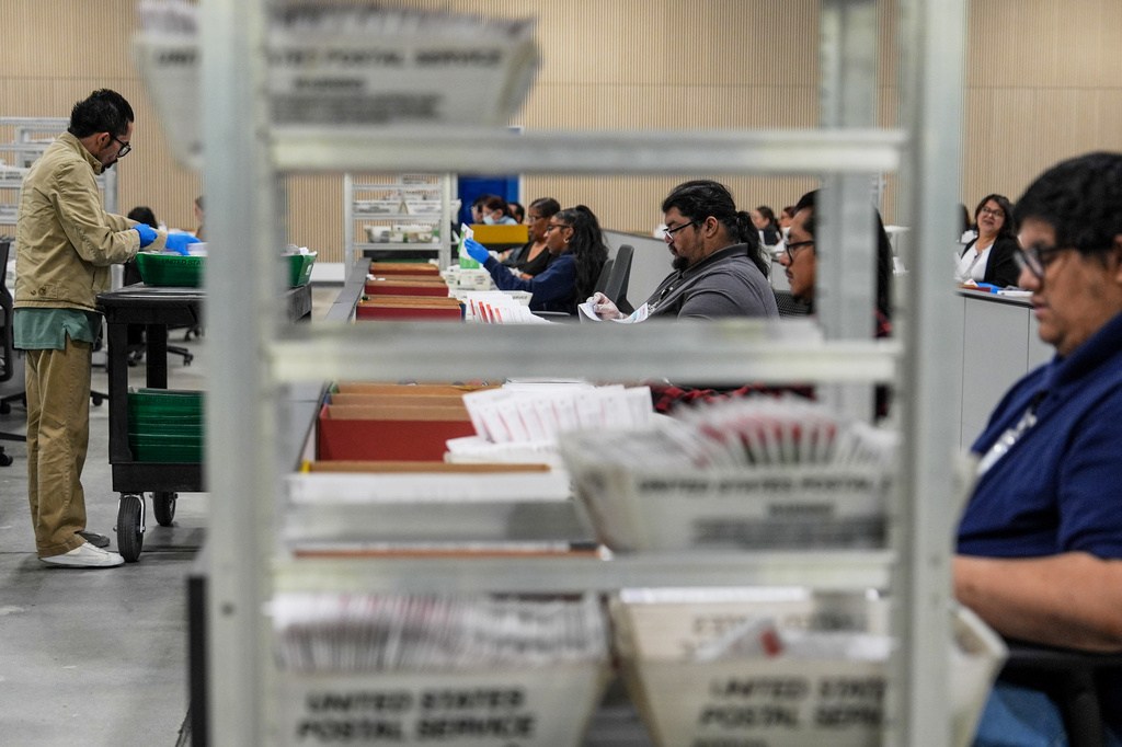 Workers handle envelopes at long tables, with trays and mail bins visible through a large rolling rack in the foreground.